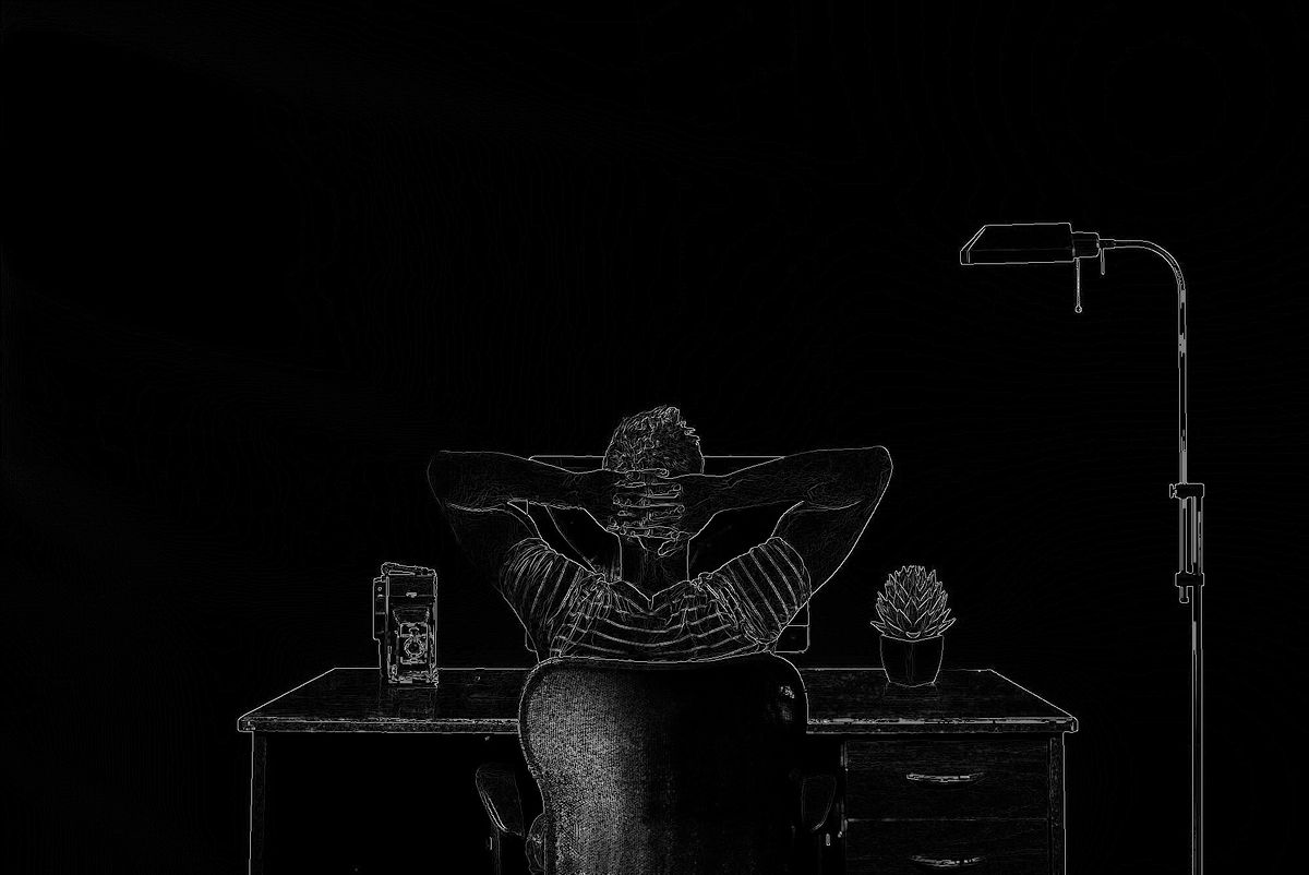 Man sitting at a desk with his hands behind his head. His back is facing you. Floor lamp to the right.